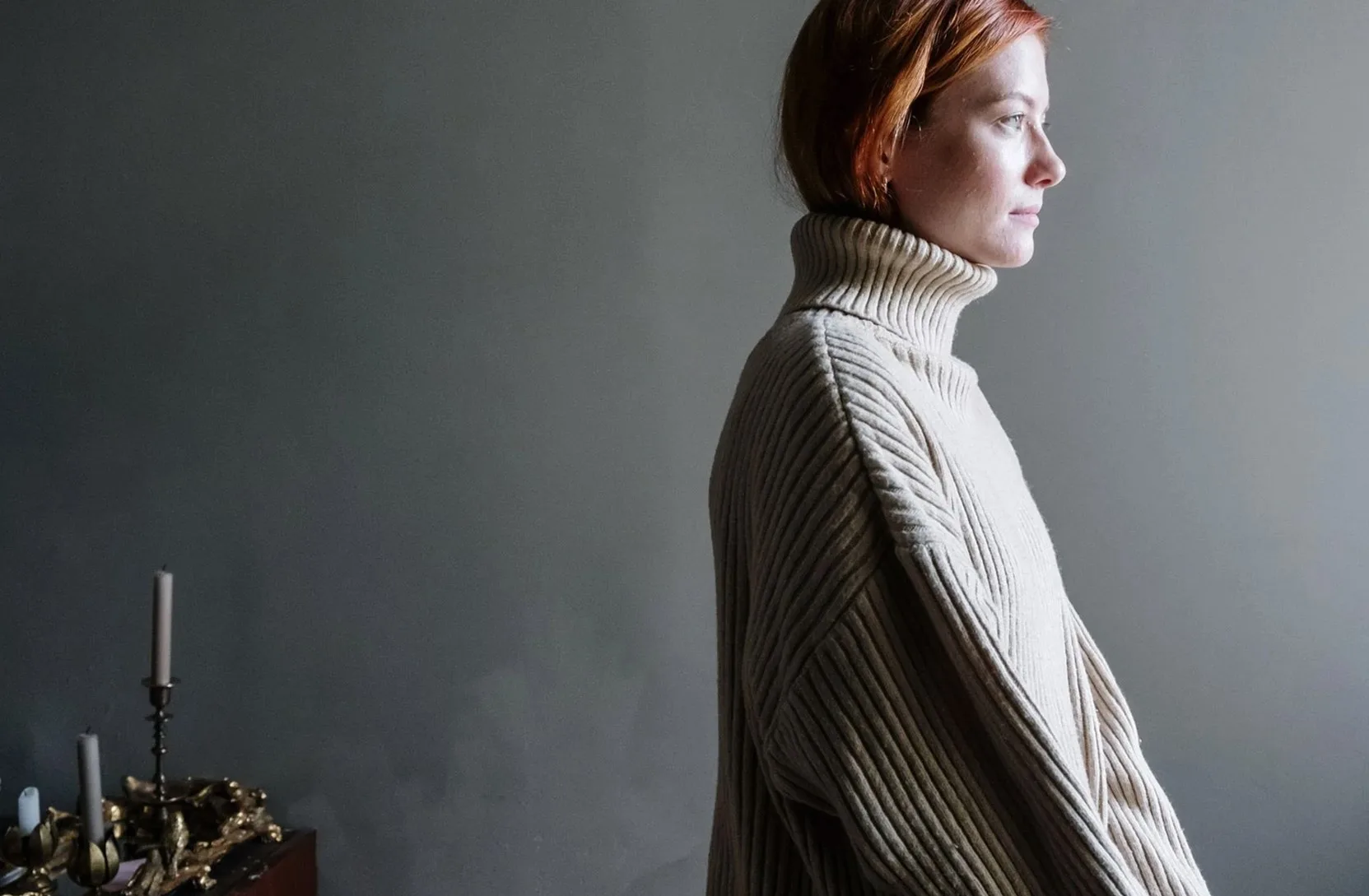 Woman in a cream ribbed turtleneck seen in profile against a dark grey wall