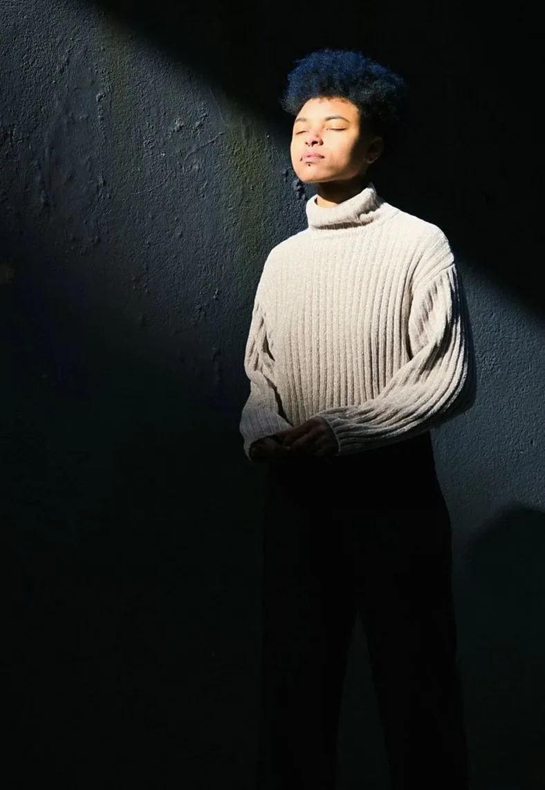 Man in a cream chunky-knit turtleneck standing in a shaft of light against a dark wall