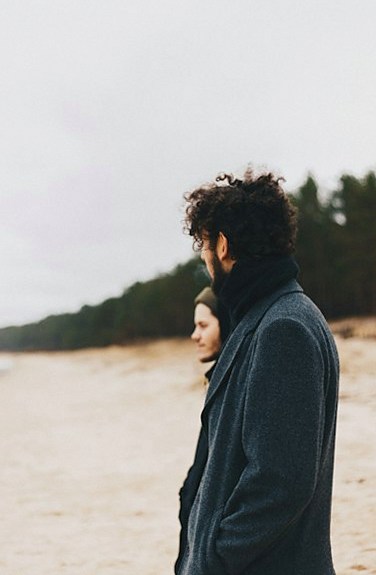 Man in a grey wool overcoat standing on a beach beside a woman