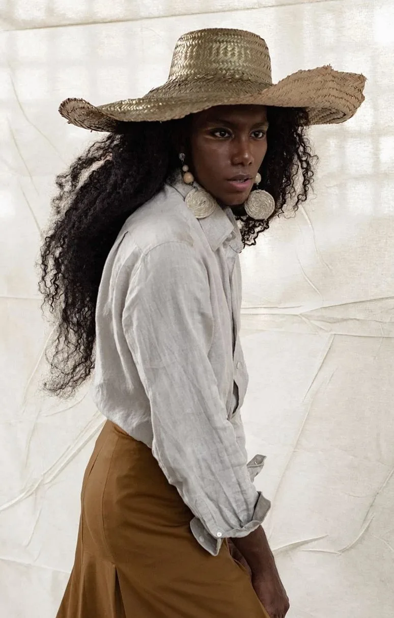 Woman in a pale linen shirt and straw hat standing against a linen backdrop